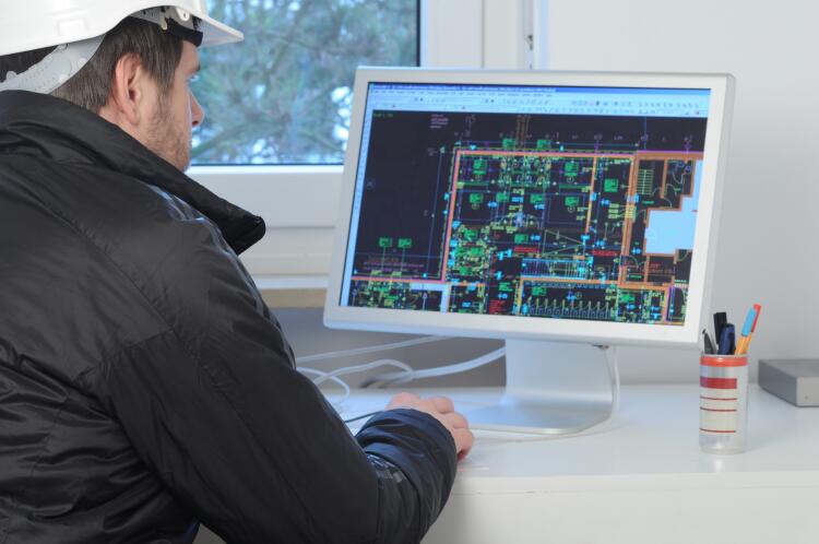 A construction engineer is seated at a desk, closely examining a detailed floor plan displayed on a computer screen. The plan includes various system layouts, highlighting the integration of Flowflex plumbing fittings and valves in a building's infrastructure.