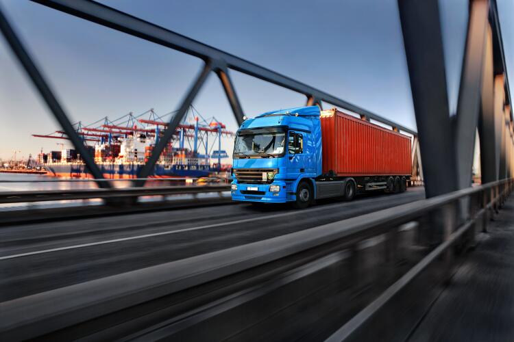 A blue lorry carrying a red container crosses a bridge with a port and cargo ships in the background. This illustrates the role of Flowflex fittings and valves in the logistics and transportation industry, ensuring the smooth operation of transport networks.