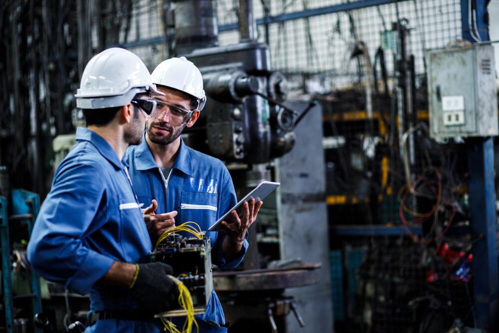 Two engineers in blue uniforms and white hard hats are discussing work inside a factory, with one holding an electronic device. This scene illustrates the collaborative and technologically advanced work environment at Flowflex, where new talent can thrive.